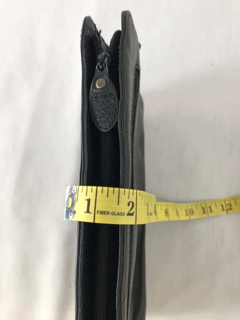 May include: Black leather clutch with a zip closure. The clutch is measured with a yellow tape measure, showing a width of approximately 5 cm.