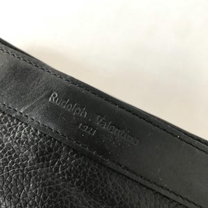 May include: Close-up of a black leather item with the embossed text "Rudolph Valentino 1921". The leather has a textured appearance, and the stitching is visible along the edge.