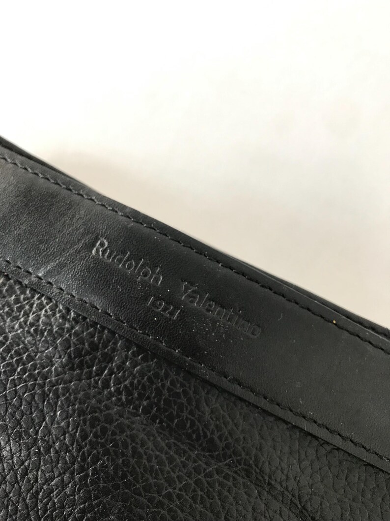 May include: Close-up of a black leather item with the embossed text "Rudolph Valentino 1921". The leather has a textured appearance, and the stitching is visible along the edge.