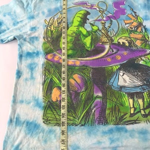Peut inclure: Un t-shirt tie-dye avec une illustration fantaisiste. Le motif pr&eacute;sente une figure verte fumant un narguil&eacute; sur un champignon, avec des &eacute;l&eacute;ments inspir&eacute;s d'Alice au pays des merveilles. Le t-shirt mesure environ 74 cm de long.