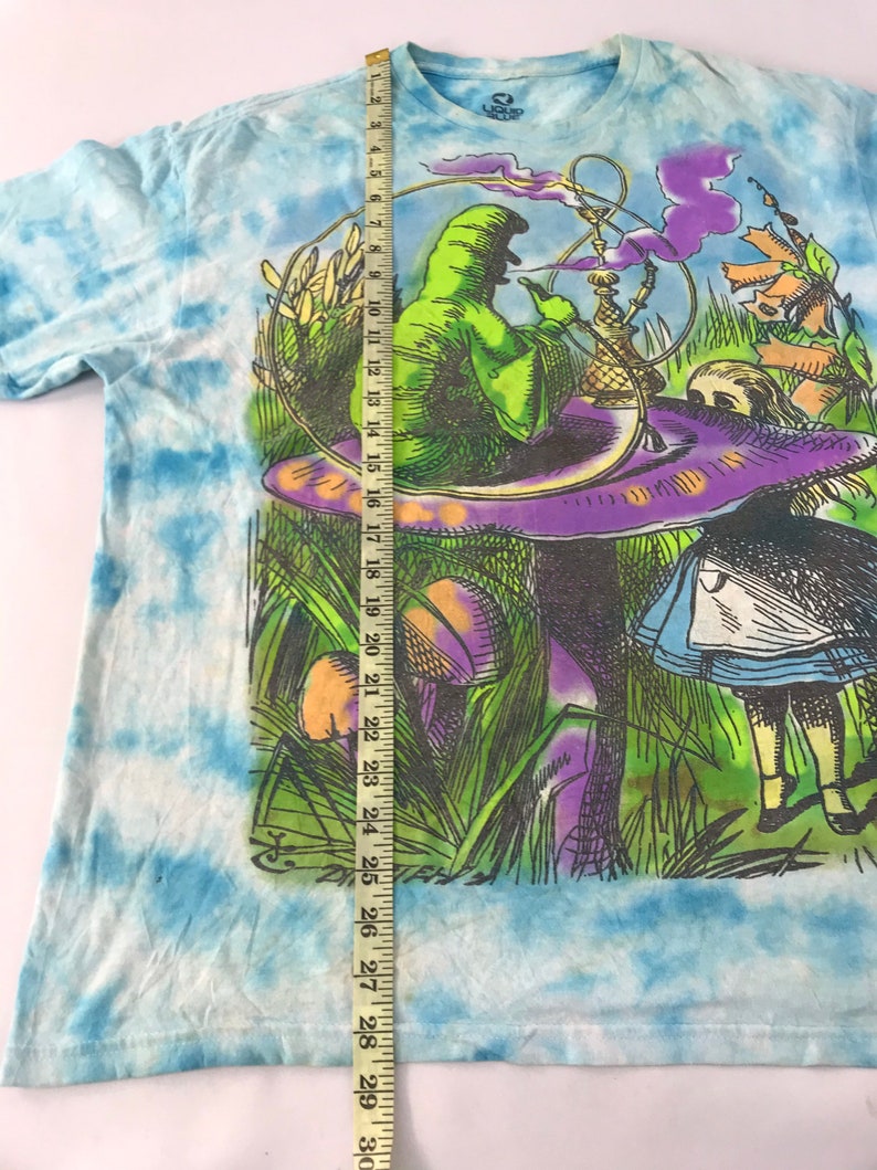 Peut inclure: Un t-shirt tie-dye avec une illustration fantaisiste. Le motif pr&eacute;sente une figure verte fumant un narguil&eacute; sur un champignon, avec des &eacute;l&eacute;ments inspir&eacute;s d'Alice au pays des merveilles. Le t-shirt mesure environ 74 cm de long.