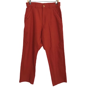 May include: Rust-coloured trousers hanging on a wooden hanger. The trousers have a button closure, belt loops, and front pockets. The fabric appears to be a medium-weight material. The trousers are a solid colour and have a relaxed fit.