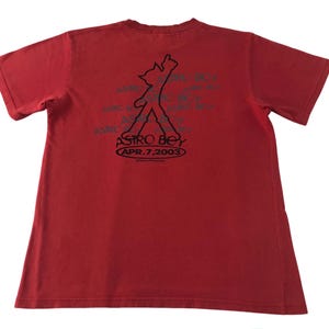 May include: Red t-shirt featuring a black graphic of a stylized figure with the text "ASTRO BOY APR. 7, 2003". The short-sleeved shirt is made of a soft material.