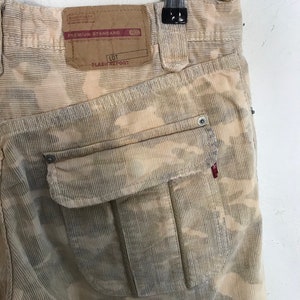 May include: Beige corduroy pants with a camouflage pattern. The pants have a large cargo pocket on the back with a white button. The pants have a label that says "Premium Standard" and "Flash Report".