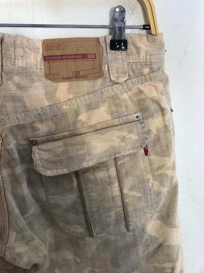 May include: Beige corduroy pants with a camouflage pattern. The pants have a large cargo pocket on the back with a white button. The pants have a label that says "Premium Standard" and "Flash Report".