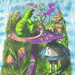 Peut inclure: T-shirt tie-dye avec un graphisme color&eacute; d'Alice au pays des merveilles. Le motif repr&eacute;sente une chenille verte fumant un narguil&eacute; sur un champignon, avec Alice au premier plan. Le t-shirt a un motif tie-dye bleu et blanc.