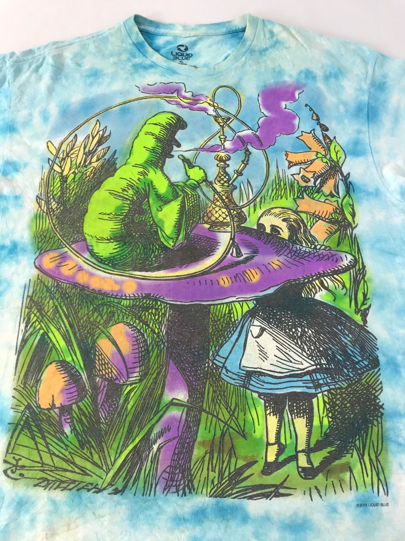 Peut inclure: T-shirt tie-dye avec un graphisme color&eacute; d'Alice au pays des merveilles. Le motif repr&eacute;sente une chenille verte fumant un narguil&eacute; sur un champignon, avec Alice au premier plan. Le t-shirt a un motif tie-dye bleu et blanc.