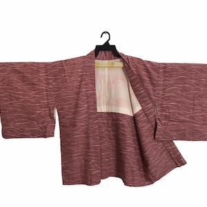 May include: A mauve-colored kimono with a wave pattern design. The kimono is open, revealing a white interior with a floral design. It is hanging on a black hanger against a white background.