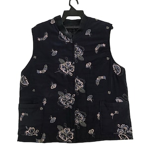 May include: A black vest with a floral pattern. The vest has two front pockets and a mandarin collar. The floral design includes flowers and butterfly shapes in shades of pink, white, and blue. The vest is sleeveless and hangs on a black hanger.
