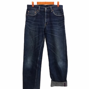 Puede incluir: Vaqueros de mezclilla azul oscuro colgados de una percha de madera. Los vaqueros tienen un diseño clásico de cinco bolsillos, bragueta con botones y dobladillo vuelto. La mezclilla tiene un aspecto desgastado con decoloración sutil.