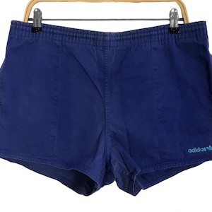 Op de afbeelding: Marineblauwe sportbroek met elastische tailleband, hangend aan een hanger. De short heeft een klein turquoise Adidas-logo op de rechter onderbeen. De short is gemaakt van een zacht materiaal, mogelijk katoen.