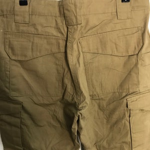 Pu&ograve; includere: Pantaloni cargo color cachi con pi&ugrave; tasche, appesi a un gancio. I pantaloni hanno una trama strutturata e un design utilitario. La vista posteriore mostra le tasche e i passanti per la cintura.