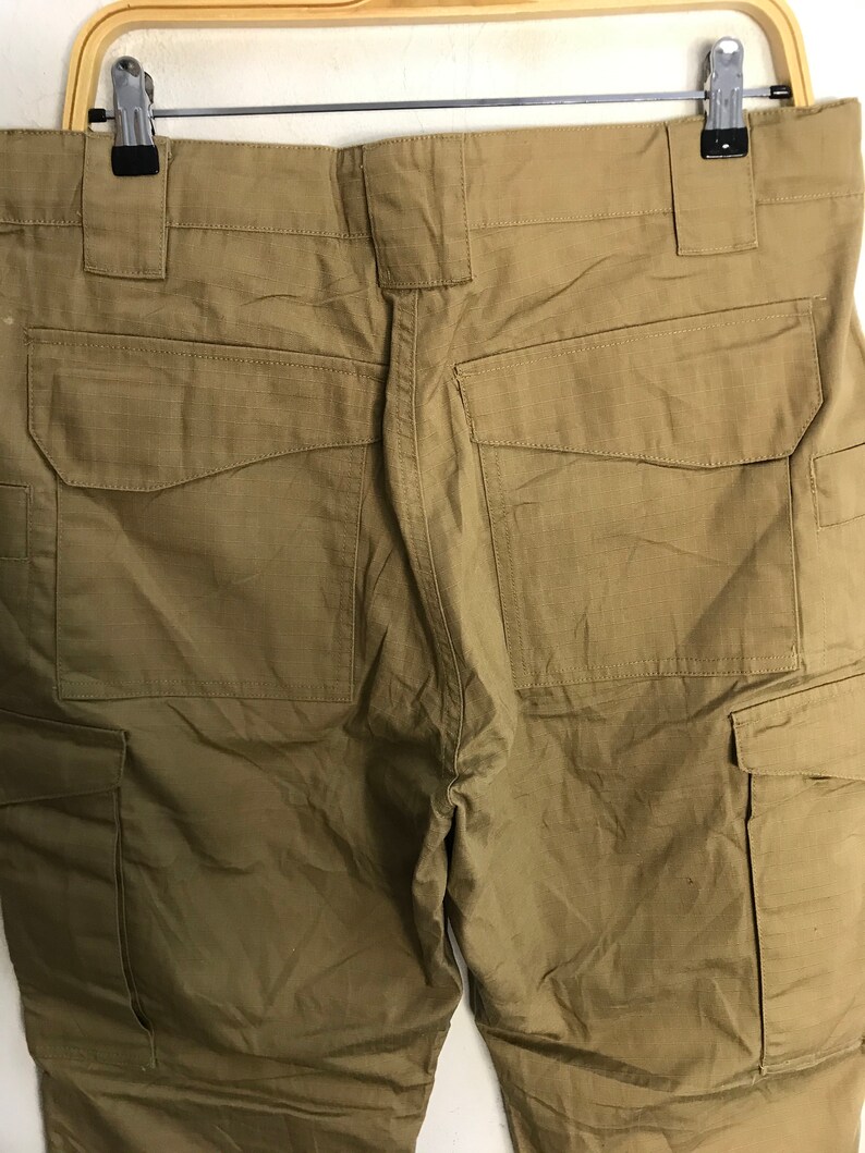 Pu&ograve; includere: Pantaloni cargo color cachi con pi&ugrave; tasche, appesi a un gancio. I pantaloni hanno una trama strutturata e un design utilitario. La vista posteriore mostra le tasche e i passanti per la cintura.