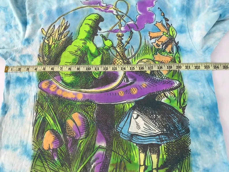 Peut inclure: Un t-shirt tie-dye avec un graphisme color&eacute; d'Alice au pays des merveilles. Le motif repr&eacute;sente une chenille verte fumant une pipe &agrave; eau sur un champignon violet, avec Alice en arri&egrave;re-plan. Le t-shirt mesure environ 63,5 cm de large.