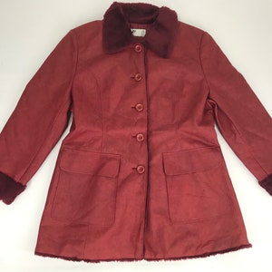 Puede incluir: Un abrigo burdeos con cuello y puños de piel sintética. El abrigo tiene una botonadura frontal, dos bolsillos delanteros y una forma ligeramente acampanada. La tela tiene una textura similar a la gamuza.