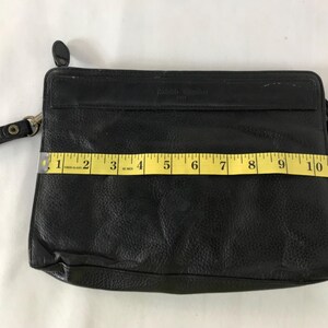 May include: Black leather clutch bag with a wrist strap. The rectangular bag is approximately 28 cm wide. The leather has a textured appearance. It features a zip closure and a small, round, black accent on the top.