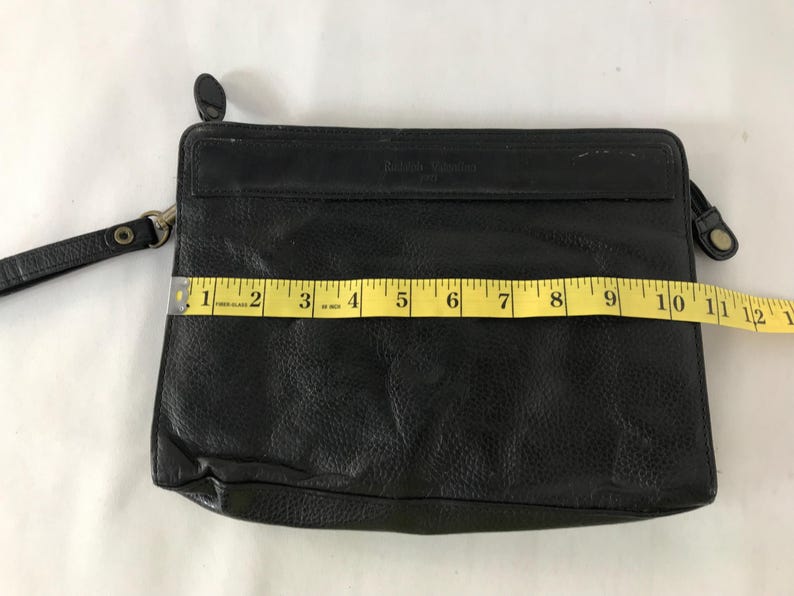 May include: Black leather clutch bag with a wrist strap. The rectangular bag is approximately 28 cm wide. The leather has a textured appearance. It features a zip closure and a small, round, black accent on the top.