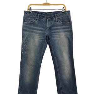 May include: Light blue denim jeans with a decorative white embroidered design on the left leg. The jeans are hanging on a wooden hanger against a white background. The jeans have a classic five-pocket design.