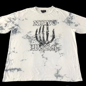 以下が含まれることがあります： 白のタイダイ染めTシャツに、黒い骨の手のグラフィックとゴシック体の「NEXT WISH」の文字がプリントされています。