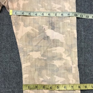 May include: A pair of beige corduroy pants with a camouflage pattern. The pants have a slight sheen and are measured with a tape measure.