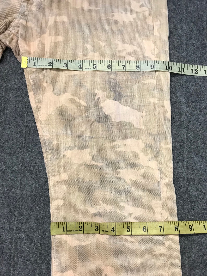 May include: A pair of beige corduroy pants with a camouflage pattern. The pants have a slight sheen and are measured with a tape measure.