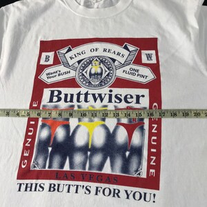 May include: White t-shirt with a graphic parody of a beer label, featuring the word "Buttwiser" and images of three posteriors in colorful underwear. The text includes "King of Rears" and "This Butt's For You!"