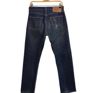 May include: Dark wash denim jeans hanging on a wooden hanger. The jeans have a classic five-pocket design, with a faded appearance and a leather patch on the back. The back pockets have a distinctive stitching pattern.