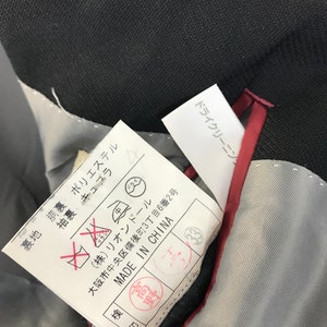 Puede incluir: Primer plano del interior de una prenda negra, que revela un forro gris y un ribete rojo. Una etiqueta blanca con texto japon&eacute;s y s&iacute;mbolos de cuidado es visible, junto con una etiqueta "MADE IN CHINA". La tela de la prenda parece ser oscura.