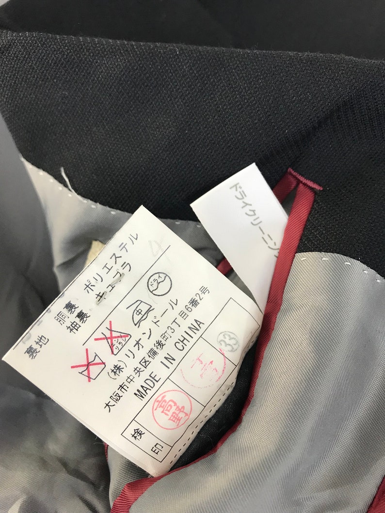 Puede incluir: Primer plano del interior de una prenda negra, que revela un forro gris y un ribete rojo. Una etiqueta blanca con texto japon&eacute;s y s&iacute;mbolos de cuidado es visible, junto con una etiqueta "MADE IN CHINA". La tela de la prenda parece ser oscura.