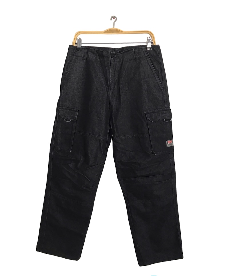 May include: Black denim cargo trousers with multiple pockets and a relaxed fit. The trousers are hanging on a wooden hanger against a white background. Features include two large side pockets and a small logo patch.
