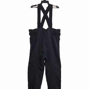 May include: Black ski trousers with braces, displayed on a black hanger. The trousers feature a high waist and adjustable braces that cross at the back. The material appears to be water-resistant.