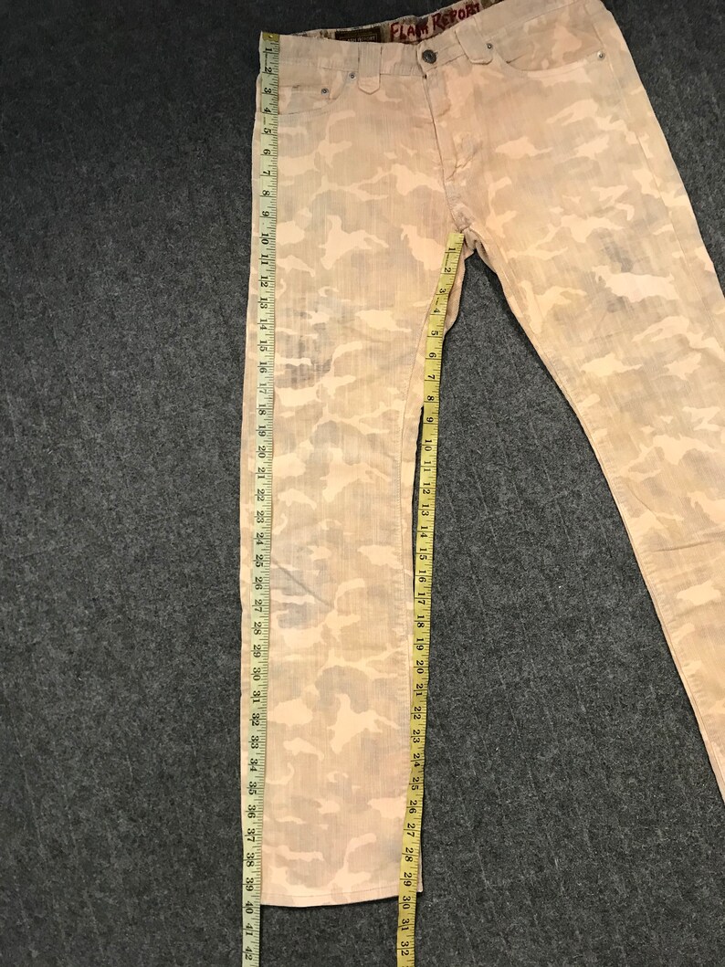 May include: A pair of light brown camouflage print jeans with a brown leather patch that says "FLAM REPORT" on the back waistband.