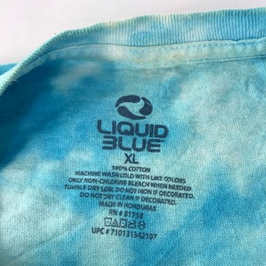 Peut inclure: Gros plan sur un t-shirt tie-dye bleu et blanc avec le logo Liquid Blue et la taille XL. Le t-shirt est 100% coton et porte des instructions de lavage. Fabriqu&eacute; au Honduras.