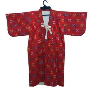 May include: A red kimono with a repeating floral pattern in blue, yellow, and pink. The kimono has wide sleeves and a white lining. A white tie closure is at the front. The garment is hanging on a black hanger.