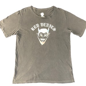 May include: A gray t-shirt featuring a devil graphic with the text "RED DEVILS" in white. The devil graphic has a red face, yellow eyes, and white horns. The shirt is a crew neck and has short sleeves.