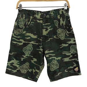 May include: Camouflage shorts in shades of green and brown, featuring a repeating pattern of stylized face designs. The shorts have a button closure, pockets, and a small patch with a cartoon character. The shorts are hanging on a wooden hanger.