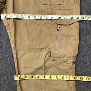 Pu&ograve; includere: Pantaloni cargo color cachi con pi&ugrave; tasche e un dettaglio con cerniera. I pantaloni sono misurati con un metro a nastro, mostrando una larghezza di circa 35.5 cm. Il tessuto ha un sottile motivo a griglia.