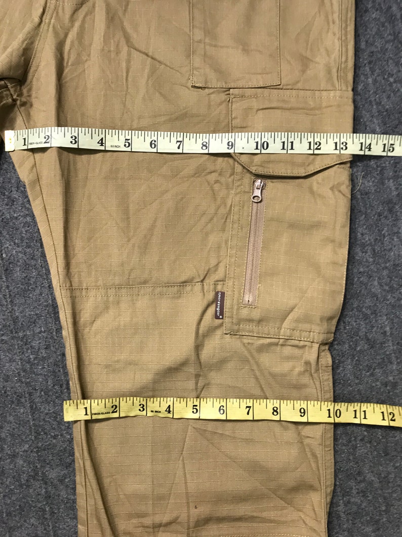 Pu&ograve; includere: Pantaloni cargo color cachi con pi&ugrave; tasche e un dettaglio con cerniera. I pantaloni sono misurati con un metro a nastro, mostrando una larghezza di circa 35.5 cm. Il tessuto ha un sottile motivo a griglia.