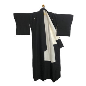 May include: A black kimono with white lining and accents, hanging on a wooden hanger. The kimono has wide sleeves and a simple, elegant design. The garment is made of a smooth fabric.