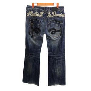 Op de afbeelding: Blauwe denim jeans met een distressed look en zilveren versieringen op de achterzakken. De achterzakken hebben de tekst "Mafia R.K. Denim" geborduurd.