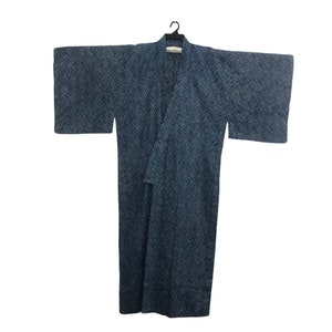 May include: A blue kimono with a textured pattern hangs from a black hanger. The kimono has wide sleeves and a wrap-around design. The fabric appears to be a dark blue with a subtle, cross-hatch pattern, creating a visual texture. The garment is displayed against a white background.