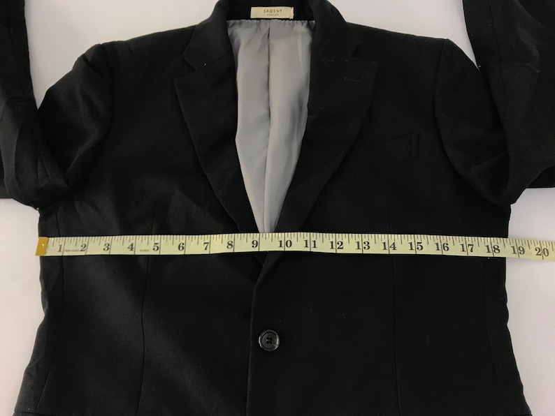 Puede incluir: Un blazer negro con cierre de un solo bot&oacute;n, solapa con muescas y forro interior gris. El blazer se mide con una cinta m&eacute;trica amarilla, que muestra un ancho de aproximadamente 50 cm.