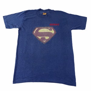 Könnte beinhalten: Marineblaues T-Shirt mit dem Superman-Logo in Diamantform und einem verblichenen Effekt. Das Wort "HENRY" ist rot über dem Logo aufgedruckt. Das T-Shirt hat kurze Ärmel und einen Rundhalsausschnitt.