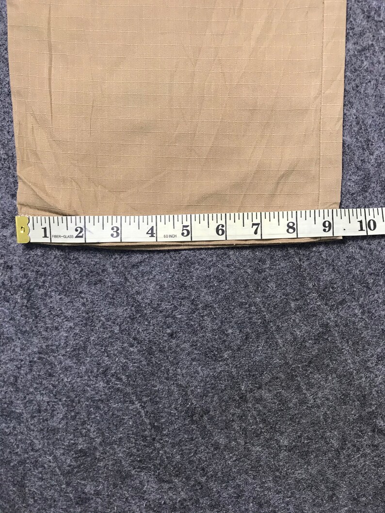 Pu&ograve; includere: Tessuto beige con motivo a griglia misurato con un metro a nastro. Il metro a nastro mostra una lunghezza di circa 25 cm. Il tessuto &egrave; su una superficie grigia.