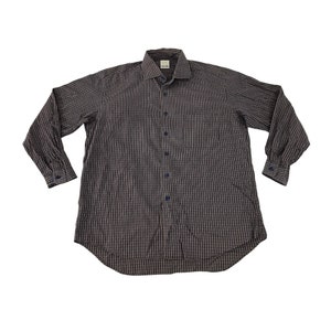 Pode incluir: Camisa de manga comprida com padrão xadrez cinza e azul. Apresenta uma gola clássica, um bolso no peito e botões azuis. As mangas também têm botões azuis.