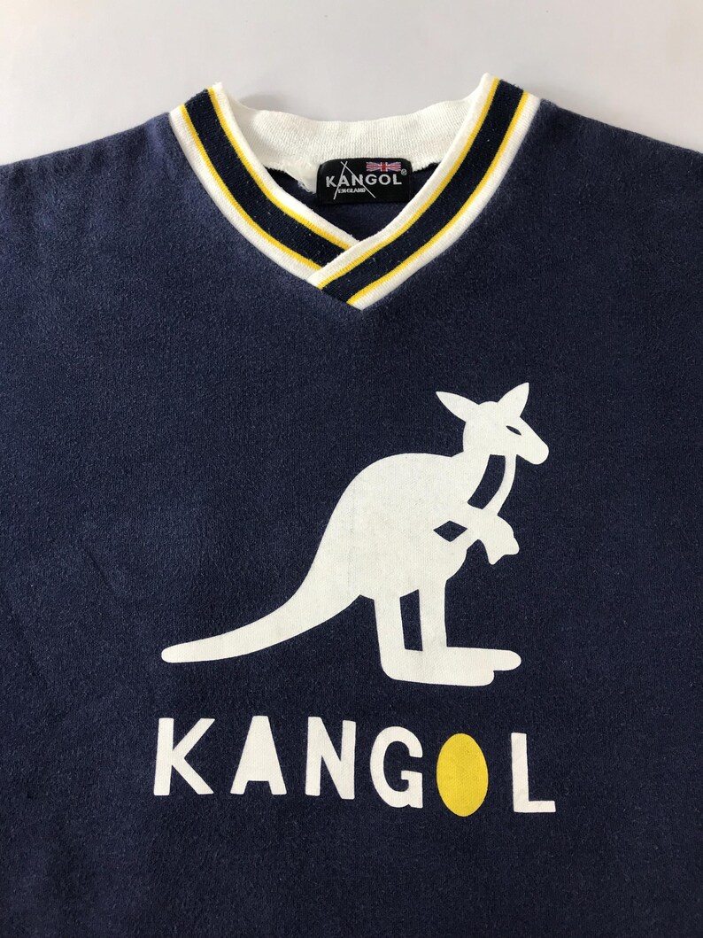 May include: Navy blue V-neck sweatshirt featuring a white kangaroo logo and the brand name "KANGOL" in white block letters. The collar has a white base with yellow and navy blue stripes. The brand's logo is visible on the collar.