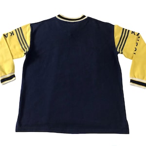 May include: Navy blue long-sleeved shirt with yellow sleeves. The sleeves have black stripes and the word "KANGOL" printed on them. The shirt has a crew neck with black and white trim.