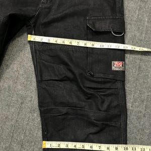 May include: Dark wash denim cargo trousers with multiple pockets and a rectangular label that reads "700". The trousers are being measured with a yellow measuring tape. The trousers are approximately 30 cm wide.