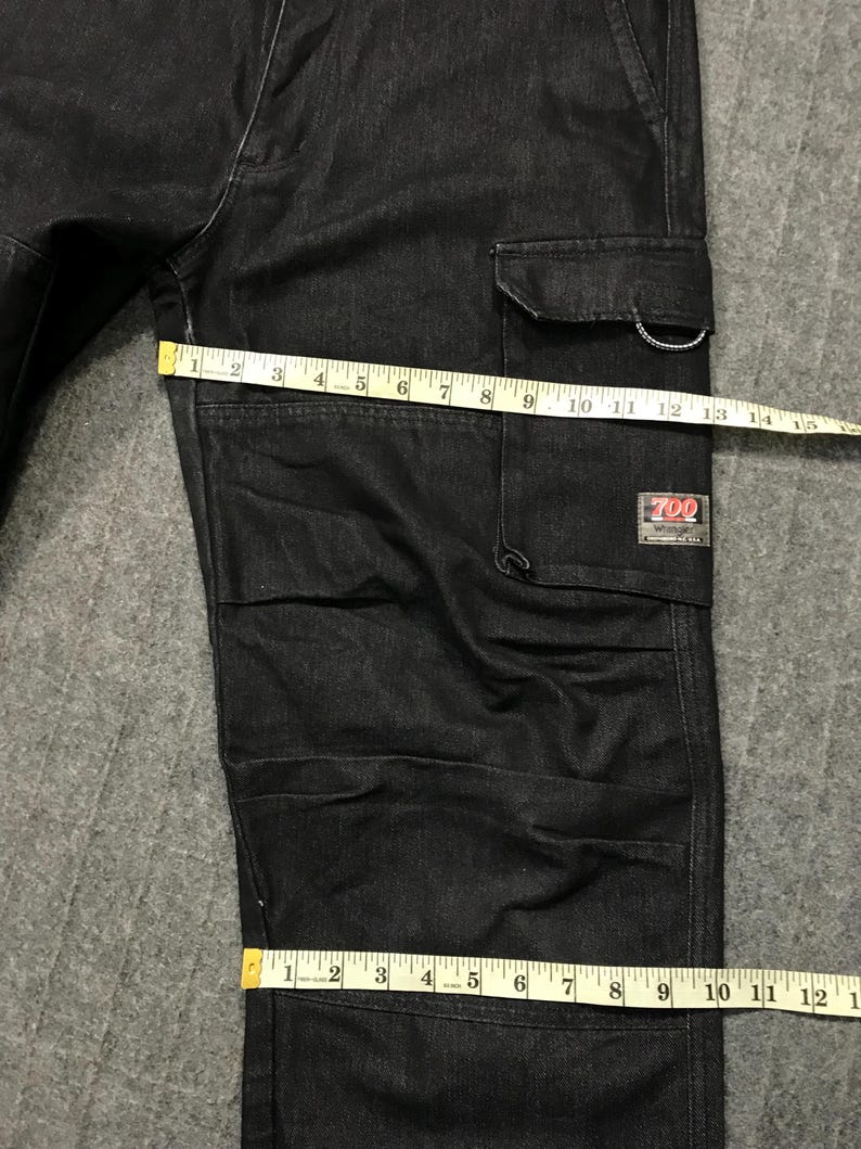 May include: Dark wash denim cargo trousers with multiple pockets and a rectangular label that reads "700". The trousers are being measured with a yellow measuring tape. The trousers are approximately 30 cm wide.
