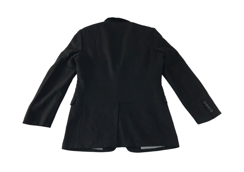 Puede incluir: Blazer negro con dise&ntilde;o cl&aacute;sico, visto desde atr&aacute;s. La chaqueta presenta solapa de muesca, dos bolsillos laterales y pu&ntilde;o con tres botones. El blazer est&aacute; confeccionado con una tela oscura y lisa.